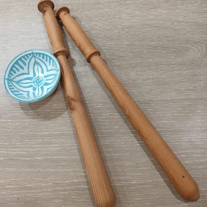 May include: Two long, light brown wooden bobbins and a small, round, white and turquoise bowl with a floral design. The bobbins are smooth and cylindrical. The bowl has a detailed, symmetrical pattern. The bobbins are likely used for lacemaking.
