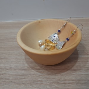 May include: A small wooden bowl with a light brown finish holds a pair of silver earrings with blue beads, a gold ring, and two white pearl earrings.