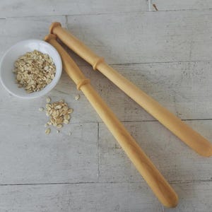 May include: Two light-coloured wooden rolling pins with a small white bowl filled with oats. A small pile of oats is next to the bowl. The items are on a light-coloured wooden surface.