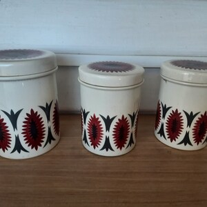 May include: Set of three white ceramic canisters with red, black and white geometric designs. The canisters are of varying sizes and are arranged in a row from largest to smallest.