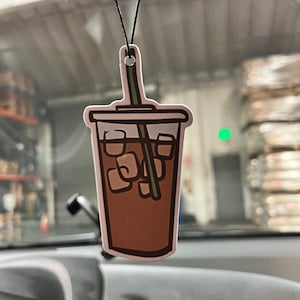 May include: A brown iced coffee drink with a green straw in a white cup with a white lid. The cup is hanging from a black string.