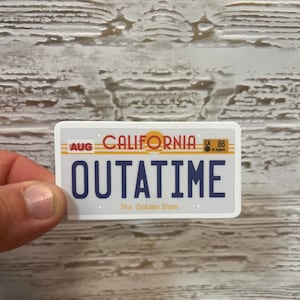 May include: A white and blue California license plate sticker with the word "OUTATIME" in blue letters. The sticker has a yellow sun graphic and the words "The Golden State" in black letters.
