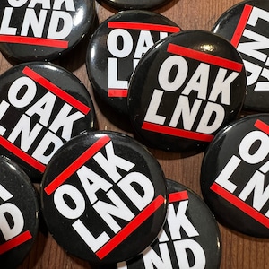 May include: A collection of black round buttons with the text "OAK LND" in white, crossed by a red line. The buttons are scattered on a wooden surface.
