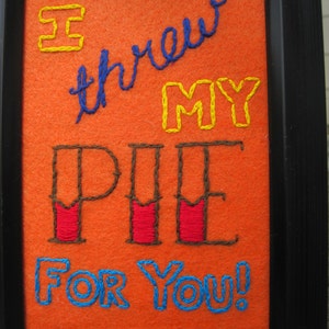 May include: Embroidered artwork on orange felt with a black frame. The artwork says "I threw my pie for you!" in colorful thread.