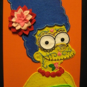 Puede incluir: Una manualidad de fieltro de un personaje de dibujos animados con cabello azul, una flor rosa y una cara amarilla con un diseño de calavera de azúcar. El personaje lleva un collar de cuentas rojas y un vestido verde.
