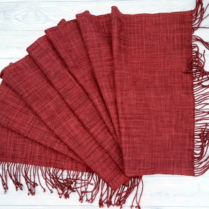 Puede incluir: Una bufanda tejida roja con flecos en los extremos. La bufanda est&aacute; doblada en forma de abanico y colocada sobre una superficie de madera blanca.