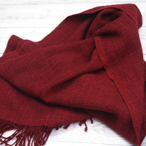Puede incluir: Una bufanda tejida de color rojo oscuro con flecos en los extremos. La bufanda est&aacute; doblada y drapeada sobre una superficie de madera blanca.