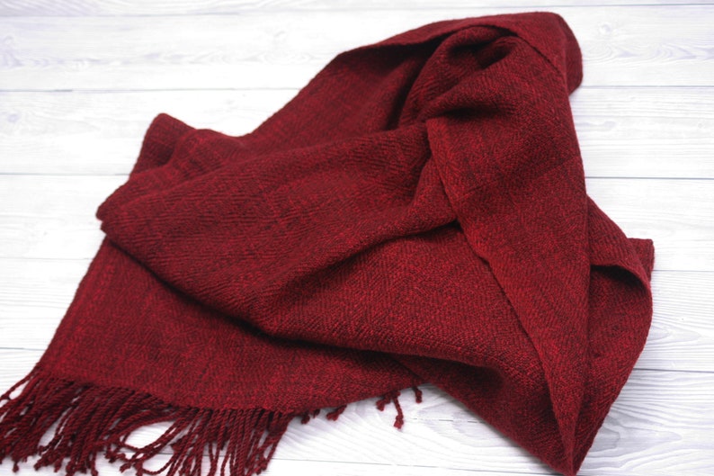 Puede incluir: Una bufanda tejida de color rojo oscuro con flecos en los extremos. La bufanda est&aacute; doblada y drapeada sobre una superficie de madera blanca.