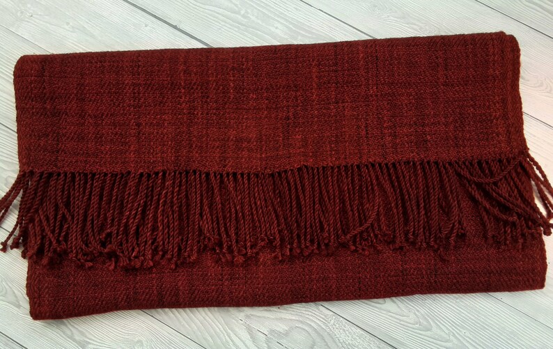 Puede incluir: Una bufanda tejida de color rojo oscuro con un borde de flecos. La bufanda est&aacute; hecha de una tela suave y texturizada.