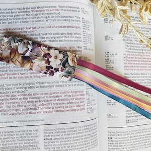 May include: A close-up of an open Bible with a floral bookmark. The bookmark is a heart-shaped fabric with a floral pattern in shades of pink, red, and white. The Bible is open to John 4:13-24, which discusses the concept of "living water" and the importance of worshipping God in spirit and truth.