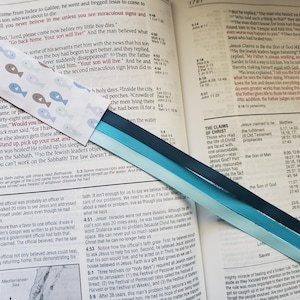 May include: A close-up of a blue, teal, and white ribbon bookmark with a pattern of fish on the white ribbon. The bookmark is placed on an open Bible with the text "The Claims of Christ" visible.