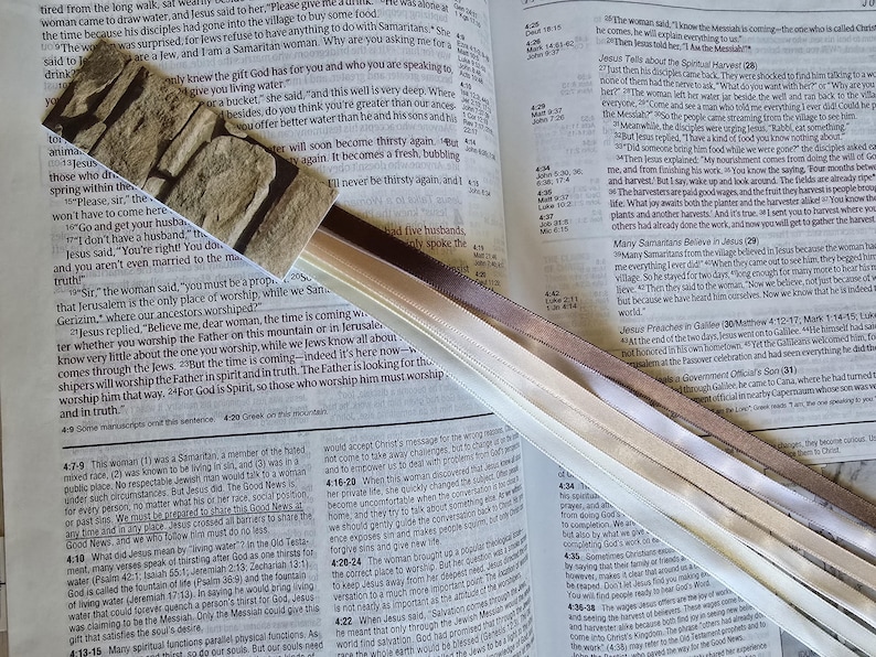 May include: A decorative bookmark with a stone-like pattern at the top and multiple ribbons in shades of brown and cream. The bookmark is placed on an open book with text, suggesting a religious or educational theme.