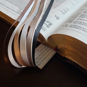 Op de afbeelding: Een close-up van een Bijbel met verschillende gekleurde linten die verschillende pagina's markeren. De linten zijn in tinten bruin, beige, wit en zwart. De Bijbel is open op een pagina met de tekst "Ories Give e.courage than Make friends and be. You have more our imagination. Ma ho you were meant" zichtbaar.