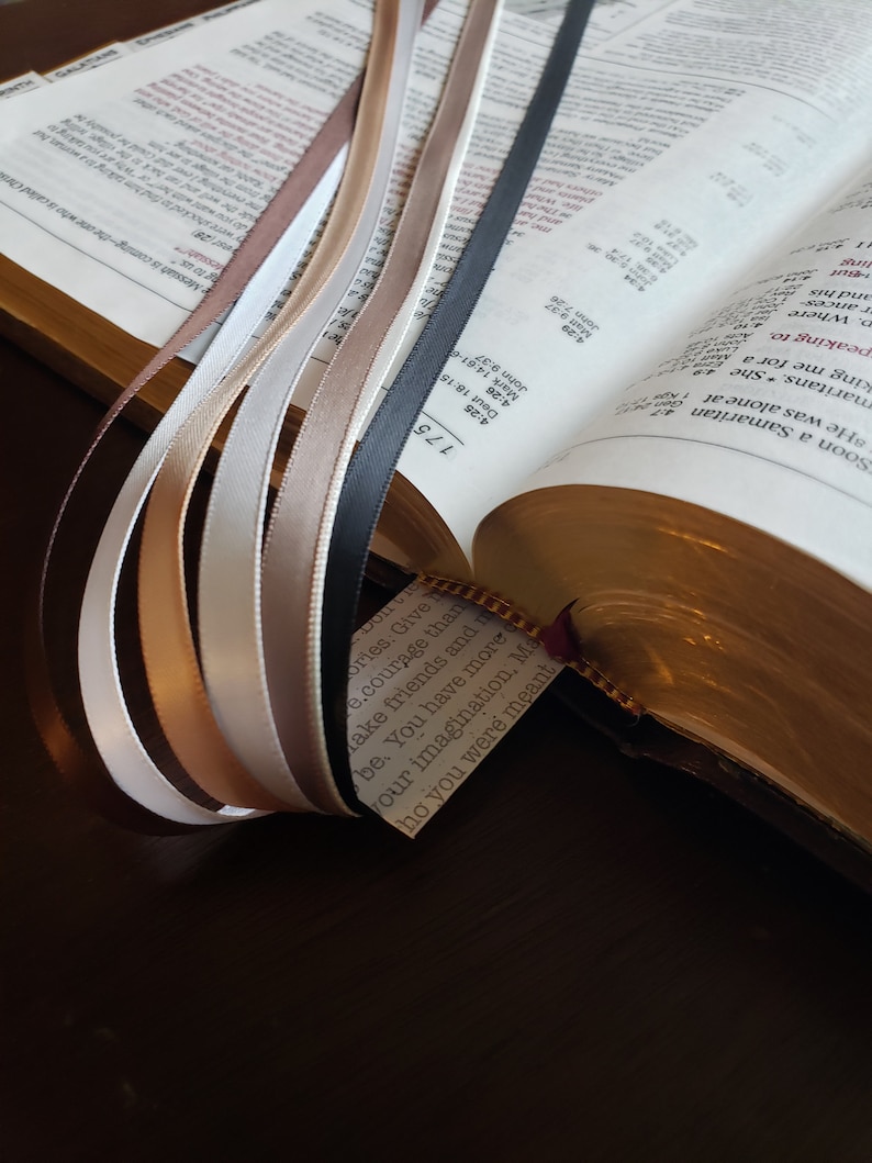 Op de afbeelding: Een close-up van een Bijbel met verschillende gekleurde linten die verschillende pagina's markeren. De linten zijn in tinten bruin, beige, wit en zwart. De Bijbel is open op een pagina met de tekst "Ories Give e.courage than Make friends and be. You have more our imagination. Ma ho you were meant" zichtbaar.