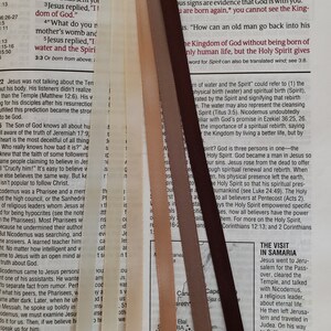 May include: A close-up of a Bible page with several colorful satin ribbons used as bookmarks. The ribbons are in shades of cream, tan, brown, and dark brown, and are placed vertically against the text.