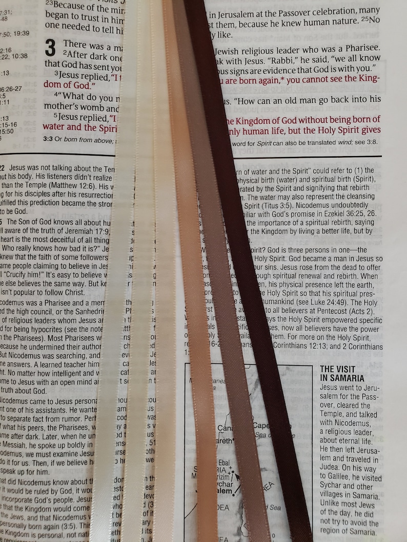 May include: A close-up of a Bible page with several colorful satin ribbons used as bookmarks. The ribbons are in shades of cream, tan, brown, and dark brown, and are placed vertically against the text.