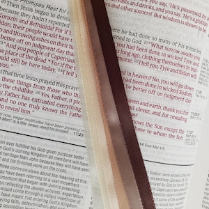 May include: Close-up of a Bible with four satin ribbon bookmarks in shades of cream, tan, and brown. The ribbons are placed between the pages of the open book, which has red text.