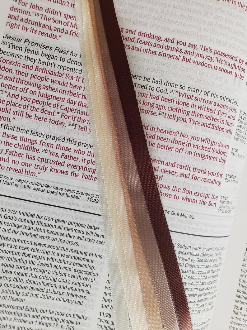 May include: Close-up of a Bible with four satin ribbon bookmarks in shades of cream, tan, and brown. The ribbons are placed between the pages of the open book, which has red text.