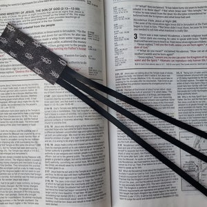May include: A close-up of a black bookmark with a white rope design. The bookmark is placed on an open Bible with black ribbon bookmarks. The Bible text is visible and includes the words "The Son of God" and "Passion for God's Authority to do".