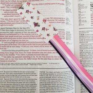 May include: A close-up of an open Bible with a pink and purple ribbon bookmark. The text on the page reads "Matthew 11:11" and continues with a passage about John the Baptist and the Kingdom of Heaven.