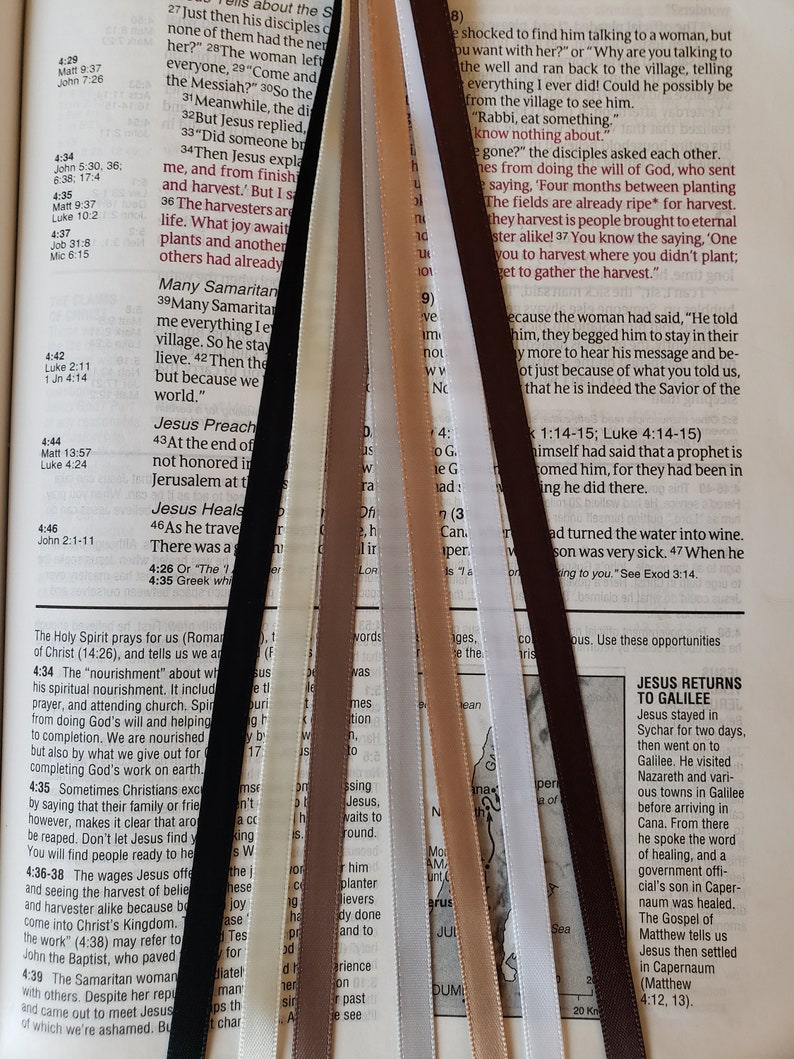 Op de afbeelding: Een close-up van een Bijbel die open ligt op het evangelie van Johannes, met verschillende gekleurde linten die verschillende verzen markeren. De linten zijn in een waaier vorm gerangschikt, met het bruine lint onderaan en het witte lint bovenaan. De tekst op de pagina bevat verzen uit het evangelie van Johannes, waaronder de verzen 4:29, 4:34, 4:35, 4:36-38, 4:39, 4:42, 4:44, 4:46 en 1:14-15. De afbeelding bevat ook een kaart van het gebied waar Jezus reisde, met de Zee van Galilea aangegeven.
