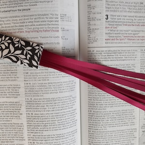 May include: A close-up of a book with a bookmark. The bookmark is made of paper with a black and white floral design. The bookmark is tucked into the pages of the book, which is open to a section about Jesus. The book is a Bible.