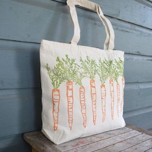 May include: A cream-colored canvas tote bag featuring a repeating screen-printed design of orange carrots with green leafy tops. The bag has two matching fabric handles attached to the top edge, providing a simple and functional carry solution.