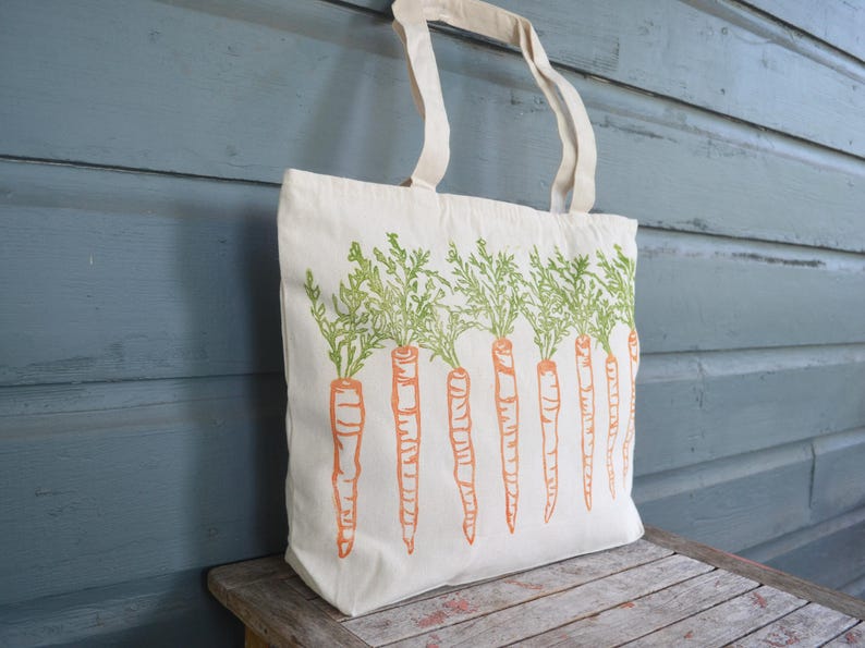 May include: A cream-colored canvas tote bag featuring a repeating screen-printed design of orange carrots with green leafy tops. The bag has two matching fabric handles attached to the top edge, providing a simple and functional carry solution.