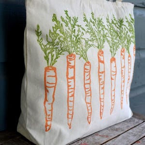 May include: A cream-colored canvas tote bag featuring a repeating block print design of orange carrots with detailed green leafy tops. The illustration is arranged in a vertical row across the front of the fabric bag, showcasing a textured ink style.