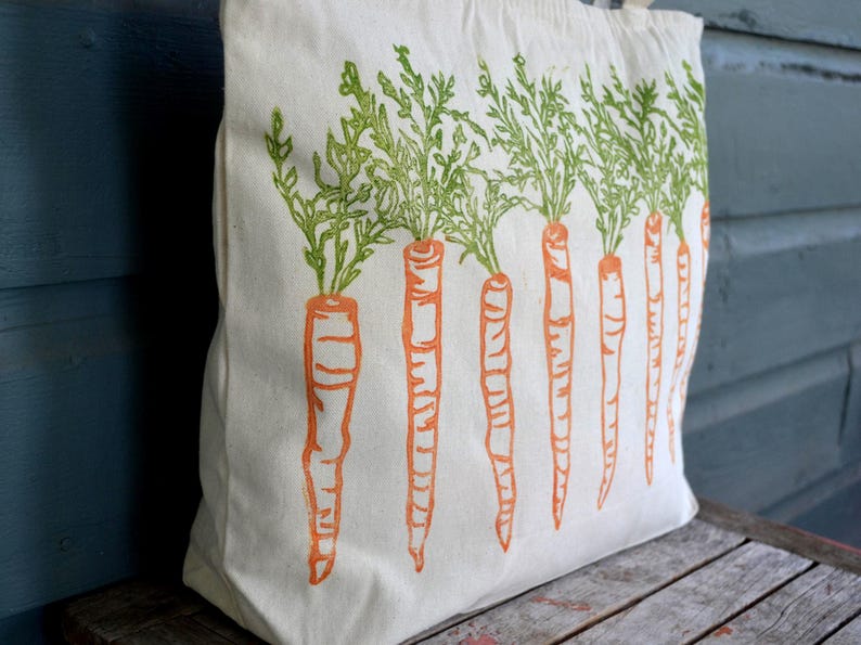 May include: A cream-colored canvas tote bag featuring a repeating block print design of orange carrots with detailed green leafy tops. The illustration is arranged in a vertical row across the front of the fabric bag, showcasing a textured ink style.