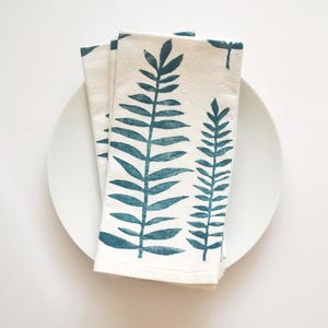 May include: A folded off-white cloth napkin featuring a dark teal botanical print of two fern-like leaves. The napkin is layered on top of a plain white circular plate, showcasing a textured fabric surface with a repeating nature-inspired pattern.