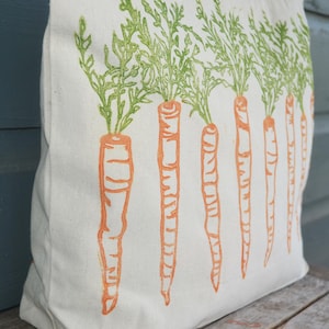 May include: A cream-colored canvas tote bag featuring a repeating block-print style design of orange carrots with vibrant green leafy tops arranged in a vertical row across the front surface of the fabric bag.