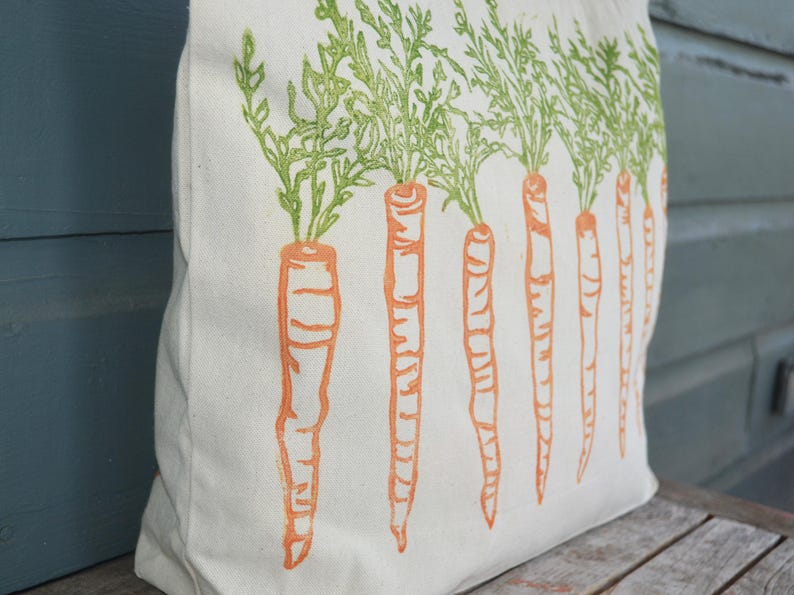 May include: A cream-colored canvas tote bag featuring a repeating block-print style design of orange carrots with vibrant green leafy tops arranged in a vertical row across the front surface of the fabric bag.
