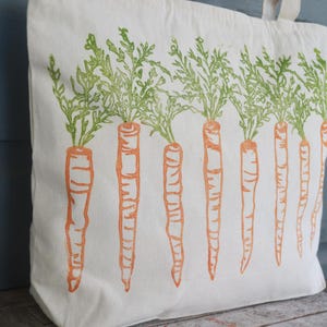 May include: A cream-colored canvas tote bag featuring a repeating block-print style pattern of orange carrots with green leafy tops arranged in a horizontal row across the front surface of the bag, which includes two fabric handles at the top.