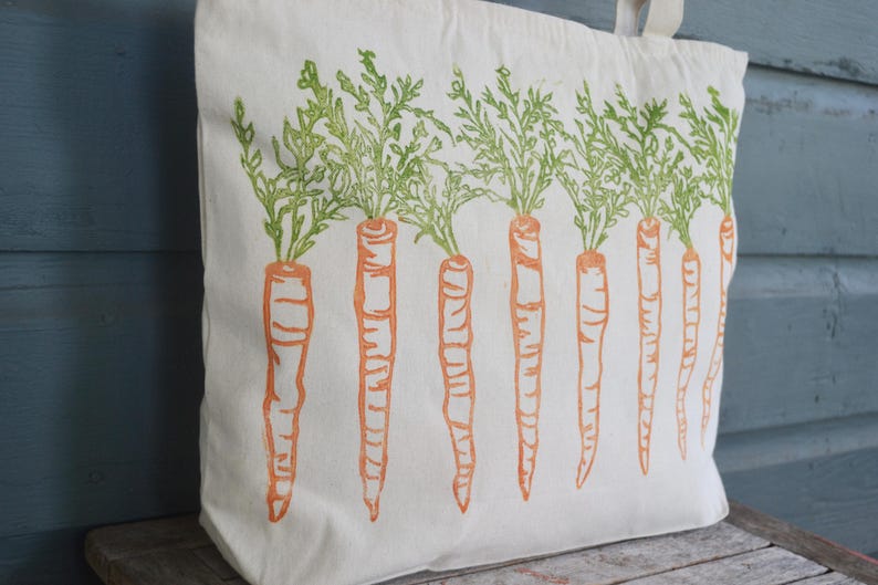 May include: A cream-colored canvas tote bag featuring a repeating block-print style pattern of orange carrots with green leafy tops arranged in a horizontal row across the front surface of the bag, which includes two fabric handles at the top.