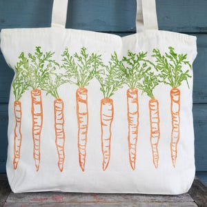 May include: A white canvas tote bag featuring a repeating screen-printed design of eight orange carrots with detailed green leafy tops arranged in a horizontal row across the front of the bag, complete with two sturdy white fabric shoulder straps.