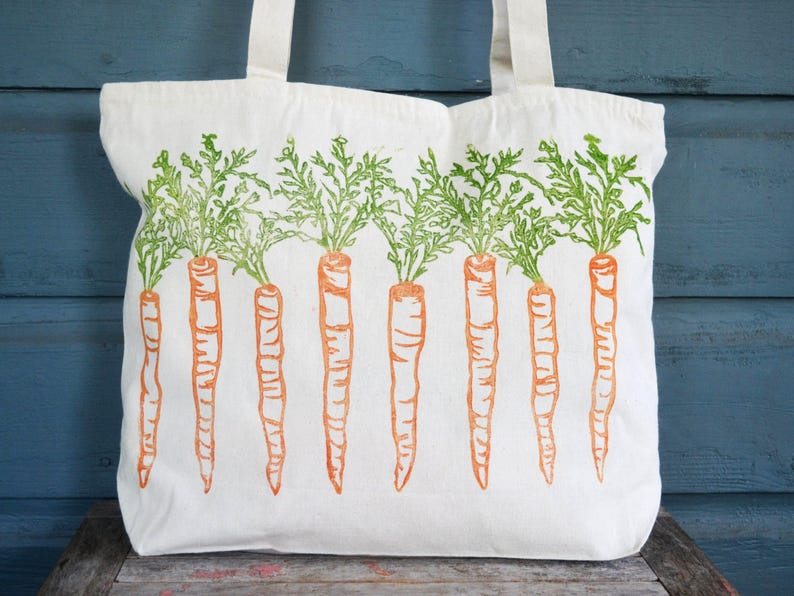 May include: A white canvas tote bag featuring a repeating screen-printed design of eight orange carrots with detailed green leafy tops arranged in a horizontal row across the front of the bag, complete with two sturdy white fabric shoulder straps.