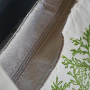 May include: A cream-colored canvas pillow cover featuring a central zipper closure. The fabric displays a printed green botanical pattern of leafy branches along the side, with a visible texture characteristic of woven cotton or linen material.
