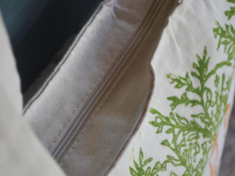 May include: A cream-colored canvas pillow cover featuring a central zipper closure. The fabric displays a printed green botanical pattern of leafy branches along the side, with a visible texture characteristic of woven cotton or linen material.