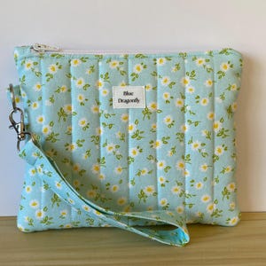 May include: A light blue quilted zipped pouch featuring a repeating pattern of small white daisies with yellow centres and green stems. A white fabric label with the text Blue Dragonfly is centred on the front, and a wrist strap is attached.