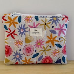 May include: A rectangular quilted zip pouch featuring a light grey background with a colourful floral pattern in shades of pink, yellow, red, blue, and purple. A white fabric label in the centre displays the text Blue Dragonfly in black.