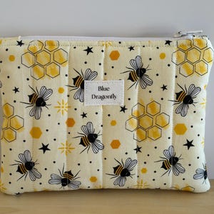 May include: A rectangular quilted pouch in pale yellow fabric featuring a repeating pattern of black and yellow bees, golden honeycombs, and small black stars. A white rectangular fabric label in the centre displays the text Blue Dragonfly.