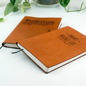 Könnte beinhalten: Zwei braune Lederjournale mit schwarzen Bandmarkierungen. Das obere Journal hat das Wort "Wanderlust" mit einer Definition auf dem Cover. Das untere Journal hat den Text "ANNAS BUCKET LIST" auf dem Cover.