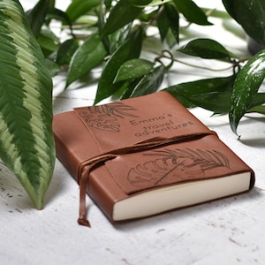 Könnte beinhalten: Ein braunes Lederjournal mit einem Lederriemenverschluss. Das Journal hat ein tropisches Blattdesign und der Text "Emma's travel adventures" ist auf dem Einband eingraviert.