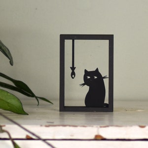 Puede incluir: Silueta negra de un gato mirando un pez colgante dentro de un marco negro. El marco está sobre una superficie blanca.