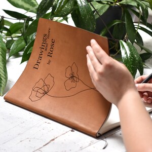 Op de afbeelding: Een bruin lederen notitieboek met de tekst "Drawings and more by Rose" en een eenvoudige lijntekening van twee bloemen op de omslag.