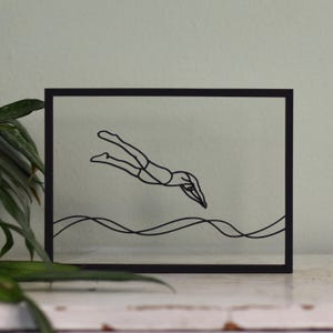 Könnte beinhalten: Schwarz gerahmtes Kunstwerk mit einer minimalistischen Strichzeichnung eines Tauchers. Das Bild zeigt eine Person, die ins Wasser taucht, mit wellenförmigen Linien, die das Wasser darstellen. Der Rahmen ist rechteckig.