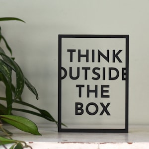 Könnte beinhalten: Schwarzes gerahmtes Schild mit dem Text "Think Outside The Box" in weißen Buchstaben.