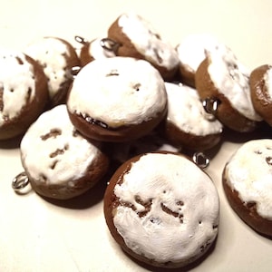 May include: A collection of miniature, round, brown and white charms. Each charm has a brown base with a white, textured top layer. Silver-colored metal loops are attached to the charms. The charms are scattered across a white surface.