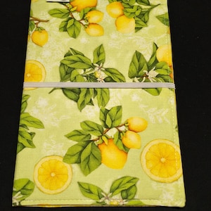 May include: A rectangular, light green fabric item with a lemon and leaf pattern. The design features yellow lemons, green leaves, and white blossoms. A gray elastic band is visible. The item is likely a journal cover or similar accessory.